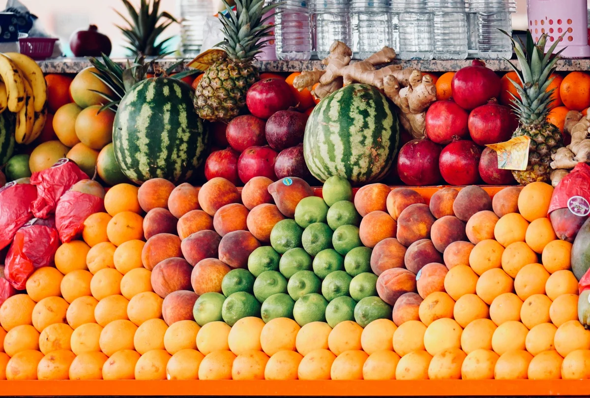 Puesto de frutería con sandías, piñas, granadas, limas y naranjas ordenadas por montones.